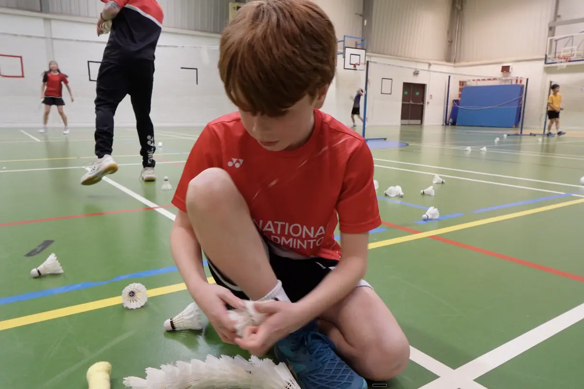 A junior player in National Badminton kit collecting shuttles on court