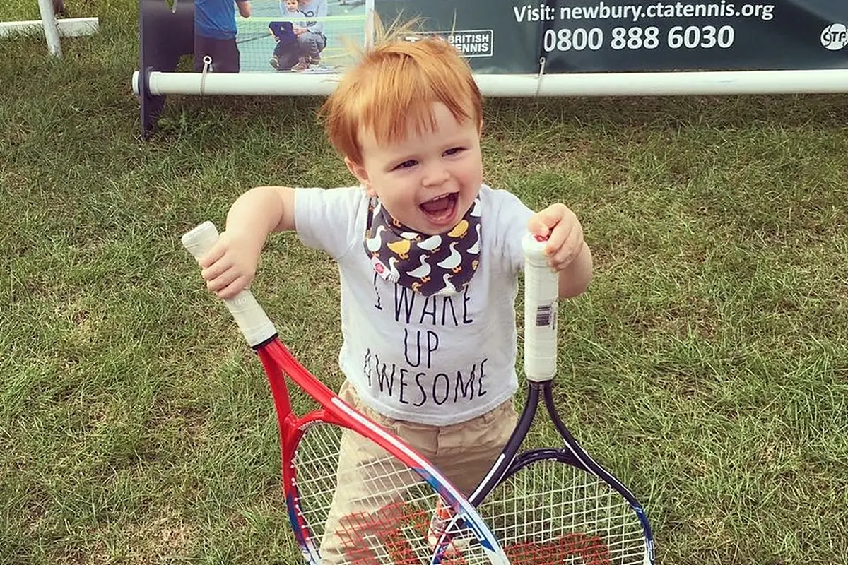 Ed’s young son holding two tennis rackets in front of a Community Tennis Association banner